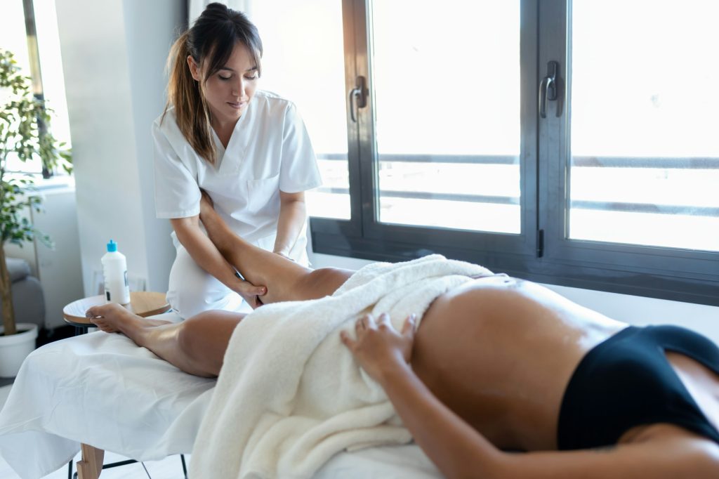 Beautiful young physiotherapist massaging the pregnant woman's legs on a stretcher at home.